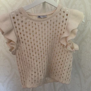 Beige stickad topp med volang Zara - Supersöt beige stickad topp från Zara med volangdetaljer på axlarna och hålmönster över hela plagget. Ärmlös modell som ger en luftig känsla och passar perfekt till både jeans och kjol. Ribbade kanter och mjukt material för en trendig look.