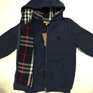 Marinblå burburry zip hoodie  - Snygg marinblå hoodie från Burberry med klassiskt rutigt foder i huvan och innerdelen. Tröjan har dragkedja, snörning i huvan och broderad logga på bröstet. Tillverkad i mjuk bomull och polyesterblandning, perfekt för kyliga dagar.