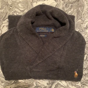 Tröja Ralph Lauren - Mörkgrå/svart  kofta från Polo Ralph Lauren med klassisk sjalkrage och broderad logga på bröstet. Tröjan är långärmad och har en mjuk, stickad känsla i bomullsmix. Perfekt för lager-på-lager och chill stil.