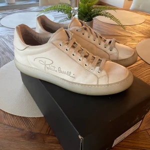 Roberto Cavalli vita skinnsneakers - Snygga vita sneakers från Roberto Cavalli i skinn med signaturbrodyr på sidan. Klassisk modell med rund tå, platt sula och snörning. Perfekta för dig som gillar stilrena och exklusiva sneakers med en touch av lyx. Lite använda hör av er vid frågor 
