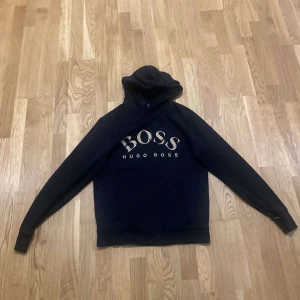Svart hoodie från Hugo Boss med guldtryck - Svart hoodie från Hugo Boss med stort BOSS-tryck i guld på bröstet. Klassisk huva med snörning och långa ärmar. Mjuk bomullskänsla och ribbade muddar. Perfekt för dig som gillar stilrena streetwear-plagg med exklusiv känsla.