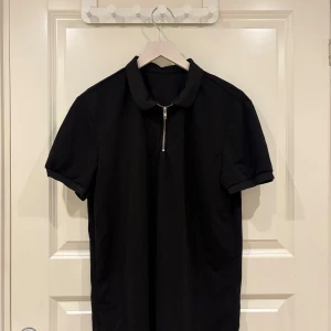 Svart piké t-shirt med dragkedja - Svart piké t-shirt med korta ärmar och klassisk krage. Framtill finns en snygg silverfärgad dragkedja som detalj. T-shirten har en stilren och enkel design, perfekt för en clean look. Materialet känns mjukt och bekvämt.