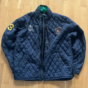 Polo Ralph Lauren jacka  - Snygg quiltad jacka från Polo Ralph Lauren i mörkblått med broderad logga och detaljer på bröstet och ärmen. Jackan har dragkedja, ståkrage och två fickor framtill. Perfekt för dig som vill ha en klassisk och sportig look. Äkta. Passar L och M. Information, bilder och prisdiskutering i dm.