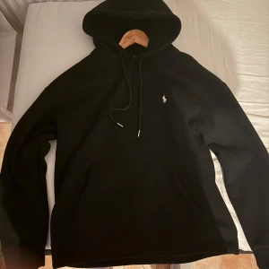 Svart hoodie från Ralph Lauren - Svart hoodie från Ralph Lauren med klassisk huva och snörning. Tröjan har en liten vit broderad logga på bröstet och en stor magficka framtill. Perfekt för dig som gillar en clean och stilren look.