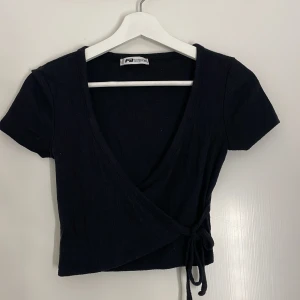 Svart omlottopp från FB Sister - Svart croppad omlottopp från FB Sister med korta ärmar och knyt i sidan. Djup v-ringning och enkel, stilren design som passar perfekt till vardagen. Mjuk och skön bomullskänsla.