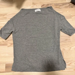 Grå t-shirt med slits från Lucky - En stilren grå t-shirt från Lucky i mjuk bomull. T-shirten har korta ärmar, rund halsringning och en liten slits i sidan för extra detalj. Perfekt basic-plagg att matcha med jeans eller kjol.
