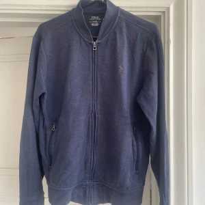 Blå ziptröja från Polo Ralph Lauren - Mörkblå långärmad ziptröja från Polo Ralph Lauren med broderad logga på bröstet. Tröjan har två fickor med dragkedja och en klassisk krage. Tillverkad i mjuk bomullsmix, perfekt för lager-på-lager-stil.