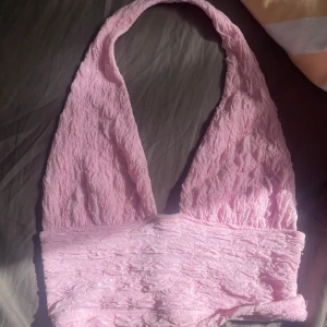 Ljusrosa omlott halterneck topp - Supersöt ljusrosa halterneck topp med omlott-design och skrynklig textur. Toppen är ärmlös och har en djup v-ringning som ger en snygg och trendig look. Perfekt att styla med jeans eller kjol för en cool vibe.