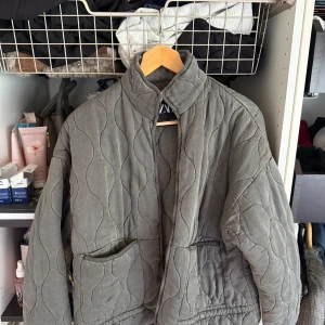 Quiltad grå jacka från ZARA - Säljer en quiltad grå jacka från ZARA i storlek M. Jackan har en hög krage, två stora fickor framtill och dragkedja. Oversized passform och vågigt mönster i tyget. Perfekt för höst och vinter. Endast använd fåtal gånger och har inga defekter. Nypris 799.