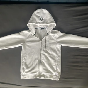 Vit hoodie från Sail Racing med dragkedja - Vit hoodie från Sail Racing med hel dragkedja framtill och en extra bröstficka med dragkedja. Tröjan har huva och långa ärmar, samt diskret logga på bröstet. Perfekt för dig som gillar stilren och sportig look.