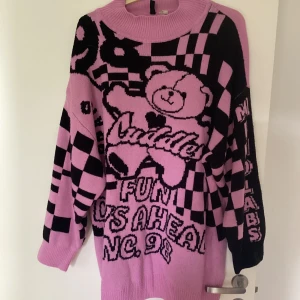 Rosa och svart stickad tröja med björn - Oversized stickad tröja  i rosa och svart med coolt rutigt mönster, stor nallebjörn och textdetaljer. Tröjan har lång ärm, hög hals och är perfekt för dig som gillar statement-plagg och lekfull stil.