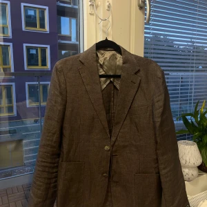 Brun linnékavaj från Oscar Jacobson - Snygg brun kavaj från Oscar Jacobson i 100% linne från Angelico. Klassisk modell med två knappar, fickor fram och slät finish. Passar storlek S/M.