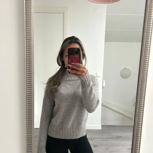 Grå tröja - Supermysig grå tröja med ribbade muddar och hög krage. Perfekt för kyliga dagar och enkel att matcha med både jeans och kjol. Tröjan har en klassisk passform och är stickad i ett mjukt material som känns skönt mot huden.