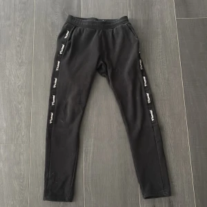 Svarta mjukisbyxor från Hummel - Svarta mjukisbyxor från Hummel med smal passform och elastisk midja. Snygga logotape-detaljer längs båda sidorna. Perfekta för chill eller träning, med mjukt material och fickor framtill.