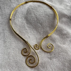 Unikt guldigt halsband med swirl-design - Säljer ett coolt halsband i guldtonad metall med snygga swirl-formade detaljer framtill. Halsbandet är stelt och ligger snyggt runt halsen, perfekt för dig som gillar statement-smycken med lite edge. Passar dig som vill sticka ut och addera något extra till din outfit.