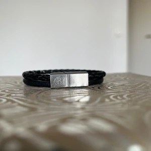 Svart flätat armband från By Billgren - Stilrent svart armband från By Billgren med dubbla band, varav ett är flätat och ett är slätt. Låset är i silverfärgad metall med logotyp ingraverad. Perfekt accessoar för dig som gillar minimalistisk och modern stil.