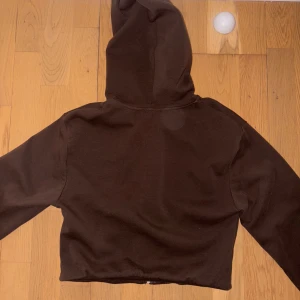 Brun croppad hoodie från Zara - Mörkbrun croppad hoodie från Zara med dragkedja framtill, snörning i huvan och en stor magficka. Tröjan har långa ärmar och är gjord i mjuk bomullsmix, perfekt för chill dagar eller när du vill ha en avslappnad look.