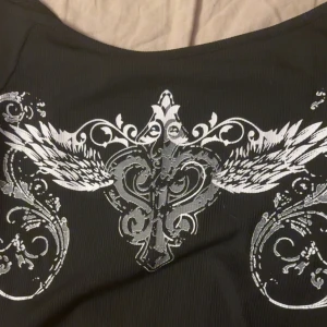 Svart långärmad topp med silvertryck - Svart långärmad topp med stort silvrigt tryck framtill som har vingar och ornament. Trycket fortsätter även på ena ärmen. Toppen har en något bredare halsringning och är figurnära i modellen. Perfekt för dig som gillar edgy och unika detaljer.