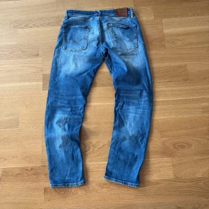 Blå jeans från Jack & Jones, 30/32 - Säljer ett par klassiska blå jeans från Jack & Jones i storlek 30/32. Jeansen har snygga slitningar och ljusare partier framtill och baktill, femficksmodell och bruna detaljer vid midjan. Perfekta för en avslappnad och trendig look. Sjuk fade