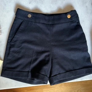 Svarta högmidjade shorts från H&M - Snygga marinblåa shorts från H&M i storlek 34 med hög midja och stilrena guldfärgade knappar framtill. 