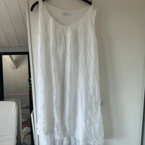 Vit långklänning Made in Italy - Luftig och flowy vit långklänning med ärmlös design och rundad hals. Klänningen har ett krinklat tyg som ger en chill vibe och är perfekt för varma dagar. Tillverkad i Italien, materialet känns som bomull eller linne. Enkel och stilren look.