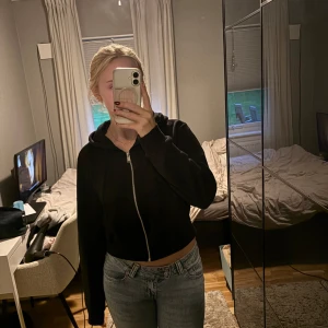 Svart croppad hoodie med dragkedja - Säljer en svart croppad hoodie med dragkedja framtill och huva. Tröjan har långa ärmar och ribbade muddar vid ärmslut och nederkant. Perfekt för en chill och avslappnad stil.