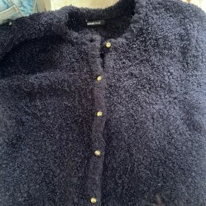 Svart fluffig kofta från Gina Tricot - Mysig svart kofta från Gina Tricot i storlek S. Koftan är fluffig och har guldiga knappar framtill. Perfekt för kyliga dagar och enkel att styla med dina favoritjeans eller kjol.