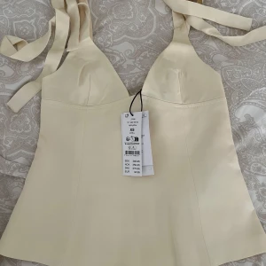 Ljusgul topp från Gina Tricot - Supersnygg ljusgul topp från Gina Tricot med  knytband vid axlarna och markerad byst. Toppen har en lätt utsvängd form och dragkedja bak för enkel påklädning. Perfekt för att skapa en chic och trendig look.