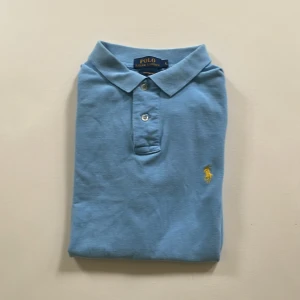 Ljusblå pikétröja från Ralph Lauren - Snygg ljusblå pikétröja från Polo Ralph Lauren i bomull. Klassisk krage, två knappar och gul broderad logga på bröstet. Custom fit-modell med korta ärmar, perfekt för en clean och stilren look. Pris ej hugget i sten!