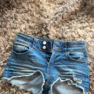 Blåa jeansshorts - Säljer ett par blåa jeansshorts som är midwaist och har fransiga kanter. Shortsen har dubbla knappar framtill, klassiska fickor och slitna detaljer för en cool look. Perfekta för varma dagar och en avslappnad stil.