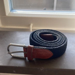 Mörkblå flätat bälte  - Snyggt mörkblått flätat bälte med detaljer i brunt skinn och ett klassiskt silverfärgat spänne. Perfekt accessoar för att lyfta din outfit med en stilren och tidlös känsla. Bältet har en robust och flexibel design som passar många stilar. Bältet är 115 cm långt.