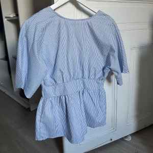 Blårandig omlottblus med knyt - Säljer en blårandig omlottblus med korta ärmar och knyt i midjan. Blusen har en luftig passform och är tillverkad i mjuk bomull. Snygg v-ringning framtill och volangdetaljer vid midjan ger en chill och trendig vibe.