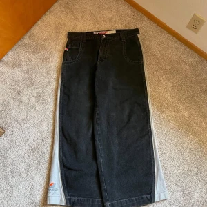 JNCO wide svarta jeans med grå sidor - Riktigt feta svarta jeans från JNCO med bred passform och grå paneler längs sidorna. Klassisk femficksmodell med coola logodetaljer på bakfickan och benet. Perfekta för dig som gillar streetwear och vill sticka ut med en oldschool vibe.