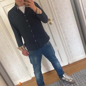 Mörkblå skjorta från Burberry - Säljer en snygg mörkblå skjorta från Burberry med klassiska rutiga detaljer på insidan av manschetterna. Skjortan har normal passform, långa ärmar och knäppning framtill. Perfekt för dig som gillar stilrena plagg. Storlek M. Säljer för 650kr. Skick 10/10 knappt avvänd, helt ny. Inget kvitto eller påse tyvärr då morsan slängde de.