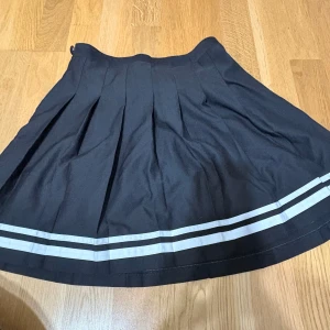 Svart plisserad kortkjol med vita ränder - Snygg svart plisserad kortkjol med två vita ränder längst ner. Kjolen har klassisk skoluniforms-vibe och är perfekt för en trendig och sportig look. Den har hög midja och är lätt utsvängd för en fin siluett.