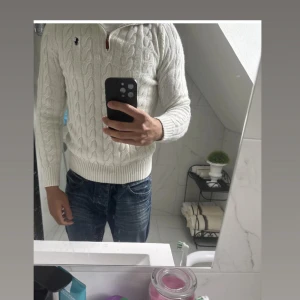 Vit stickad tröja från Ralph Lauren - Säljer en vit kabelstickad tröja från Ralph Lauren med half zip och broderad logga på bröstet. Tröjan har lång ärm, ribbade muddar och klassisk passform. Perfekt för dig som gillar stilren och tidlös look. Helt ny aldrig använd. Den passar S  