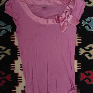 Rosa topp med rosett och satinkant - Söt rosa topp med korta ärmar och en bred satinkant runt halsen. Dekorerad med en stor rosett i satin på ena axeln för extra detalj