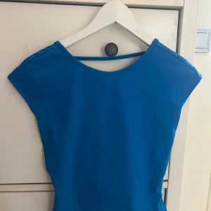 Blå kort topp med öppen rygg - Snygg blå kort topp med cool öppen rygg och breda axlar. Klänningen har en enkel, tight passform och är tillverkad i ett mjukt syntetmaterial som sitter skönt mot kroppen. Perfekt för dig som vill sticka ut med en färgstark look.