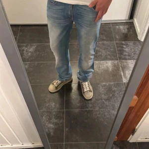 Ljusblå jeans från Nudie Jeans - Säljer ett par ljusblå jeans från Nudie Jeans med klassiska orangea sömmar och snygg broderad bakficka. Jeansen har rak passform och normal midja, tillverkade i mjuk bomullsdenim. Perfekta för dig som gillar en avslappnad och stilren look. Modellen är ”slim Jim”