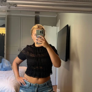 Svart spetsig croppad blus med volanger - Supersöt svart blus med kort ärm, croppad passform och massor av volanger. Hela toppen är i genombruten spets med små rynkade detaljer och hög hals. Perfekt för dig som gillar romantisk och trendig stil. Aldrig använt