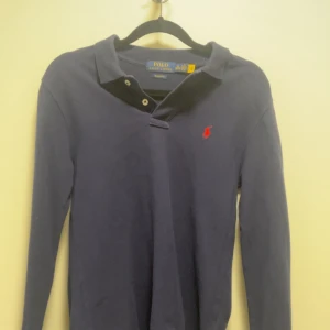 Mörkblå pikétröja från Polo Ralph Lauren - Klassisk mörkblå långärmad pikétröja från Polo Ralph Lauren med röd broderad logga på bröstet. Tröjan har krage, knappar vid halsen och är i bomull. Perfekt för en clean och tidlös stil. Bra skick. Skriv bara om frågor. Pris kan diskuteras.