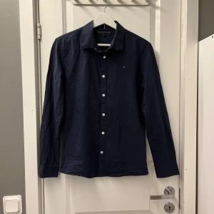 Mörkblå skjorta från Tommy Hilfiger - Snygg mörkblå skjorta från Tommy Hilfiger med klassisk krage och vita knappar. Skjortan har lång ärm och en liten broderad logga på bröstet. Tillverkad i mjuk bomull som känns skön mot huden. Perfekt för dig som gillar stilrena och tidlösa plagg. (Den är ganska stor för sin storlek passar xs)