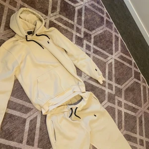 Beige hoodie från Polo Ralph Lauren - Säljer en beige hoodie från Polo Ralph Lauren med svart broderad logga på bröstet och ärmen. Tröjan har huva med svarta snören, känguruficka och är i mjuk bomull. Perfekt för chill dagar och snygg att matcha med jeans eller sweatpants.