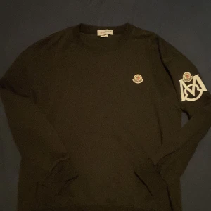Svart långärmad tröja från Moncler M/L - Svart långärmad tröja från Moncler med klassisk logga på bröstet och stor vit M-detalj samt logga på ena ärmen. Tröjan har rund hals och är i mjukt material, perfekt för dig som gillar stilrena och exklusiva plagg.