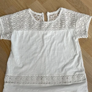 Vit spetsblus från Zara Girls - Superfin vit blus från Zara Girls med korta ärmar och detaljer i spets upptill och nertill. Blusen har ett snyggt virkat mönster på axlar och ärmar samt en liten knapp i nacken. Perfekt för dig som gillar en söt och stilren look.