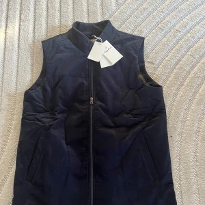 Mörkblå manchesterväst från Sayless - Snygg mörkblå dunväst från Sayless i manchester med hög krage och dragkedja framtill. Västen har en stilren look med raka linjer och två diskreta fickor på sidorna. Perfekt för lager-på-lager och passar till många outfits.