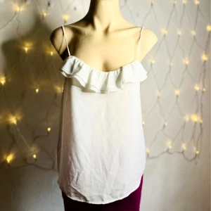 Skir vit topp med volang – stl M, somrig & feminin - Skir och lätt vit topp med tunna axelband och volang längs ringningen. Perfekt för sommarens varma dagar eller som layering-piece under kavaj eller kofta. Tillverkad i tunt vävt tyg (troligen polyester) som ger en luftig och feminin känsla. Märkning saknas men uppskattas passa storlek M. Mycket fint skick – ser knappt använd ut. Jag prutar ej, men erbjuder rabatt vid köp av flera plagg! 