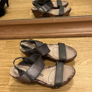 Gråa sandaler med kilklack - Säljer ett par gråa sandaler med breda remmar och kilklack. Skorna har en öppen tå och en bekväm platt sula med vadderad innersula. Sidan av klacken har ett kamouflagemönster i bruna och beige toner. Perfekta för sommaren!