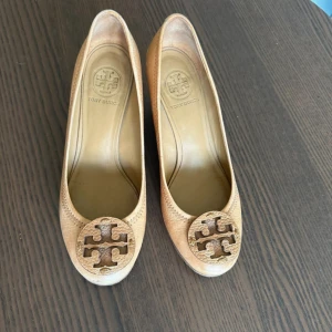 Beige kilklackar från Tory Burch - Säljer ett par snygga beige kilklackar från Tory Burch med rund tå och stort logomärke framtill. Skorna har en träfärgad kilklack och är tillverkade i skinn med struktur. Perfekta för dig som vill ha både stil och komfort.