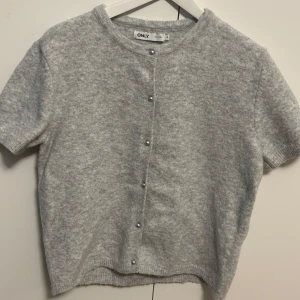 Ljusgrå kofta med pärlknappar från ONLY - Supermjuk ljusgrå kofta från ONLY med korta ärmar och rund hals. Stängs med snygga pärlknappar framtill. Perfekt att styla över en topp eller klänning. Materialet känns fluffigt och mysigt.